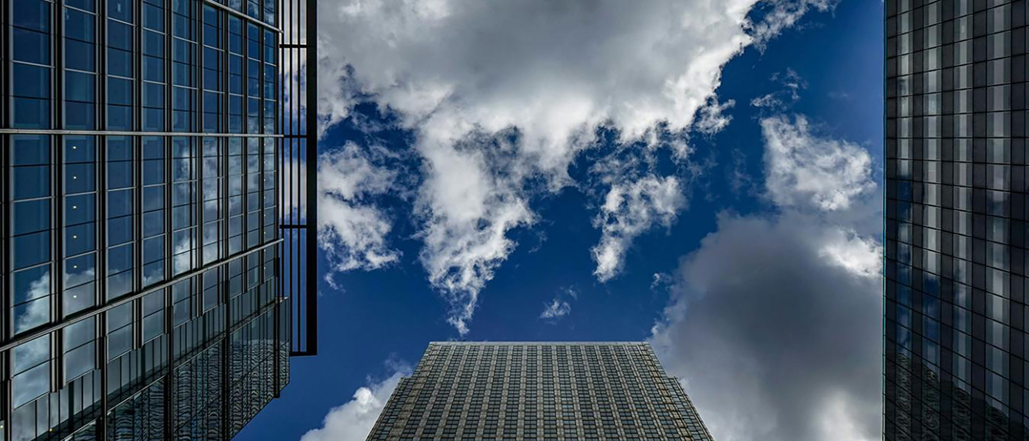 Looking up between tall glass buildings toward sky and clouds