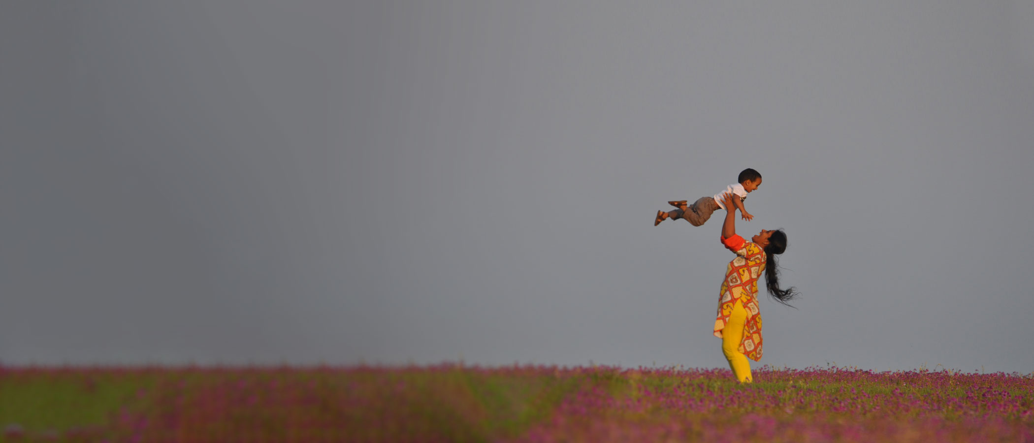 A south asian woman holding a child above her head in a field of wild flowers.
