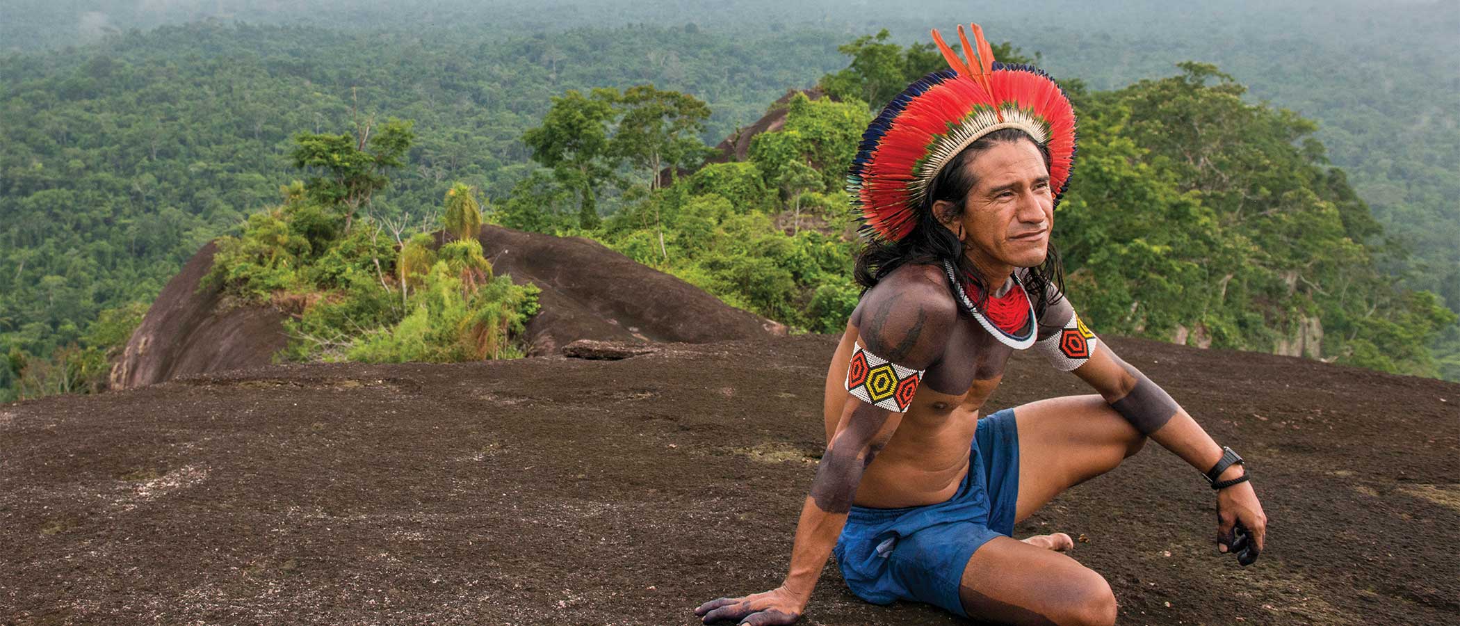 A Kayapo man seated on a mountain top overlooking forest in the Amazon.